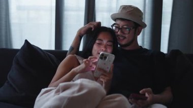 Genç Asyalı çift kanepeye uzanmış akıllı telefon kullanıyor ve hassas bir anı paylaşıyorlar - 4K Yatay