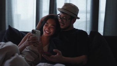 Mutlu genç Asyalı çift rahatlarken akıllı telefon kullanıyor ve birlikte dizi izliyorlar - 4K Yatay