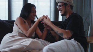 Genç Asyalı çift el ele tutuşup gülüyor ve birlikte kanepede vakit geçiriyorlar - 4K Yatay