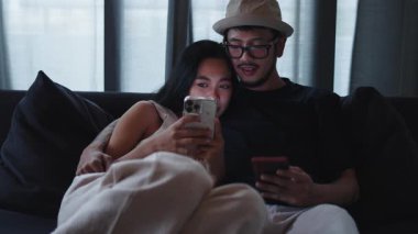 Genç ve mutlu Taylandlı çift kanepeye uzanmış akıllı telefonlarını kullanıyor - 4K Yatay