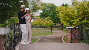 Genç Çok Irklı Çift Parktaki Köprü korkuluklarına yaslanırken Akıllı Telefon Kullanıyor - 4K Yatay