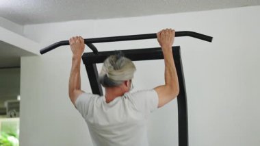 Sırtından uzun saçları olan yaşlı bir adam spor salonundaki bir barda barfiks egzersizi yapıyor - 4K Yatay