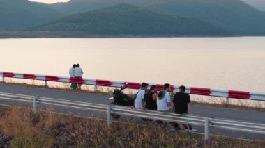 Yol kenarındaki bir bariyerde oturan arkadaşlar gölün yanında huzurlu bir manzaranın tadını çıkarıyorlar. Arka planda dağlar var. 4K Yatay