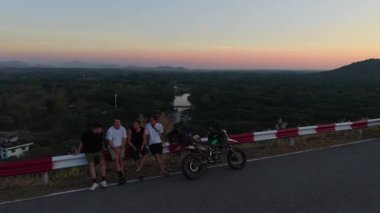Yol kenarındaki bariyerde dinlenen bir grup motorcu vadi ve nehrin üzerindeki güzel günbatımını hayranlıkla seyrediyor. 4K Yatay