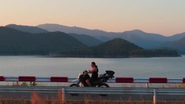 Gün batımında gölün yanında manzaralı motosiklet yolculuğunun keyfini çıkaran genç bir çocuk - 4K Yatay
