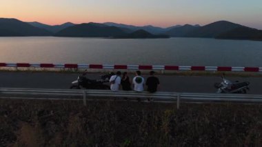 Gün batımında göl ve dağların panoramik manzarasının keyfini çıkaran bir grup arkadaş motosikletlerinin yanında - 4K Yatay