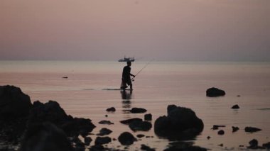 Siluet. Bir balıkçı gün batımında olta ve balık ağıyla sığ sularda yürüyor. 4K Yatay