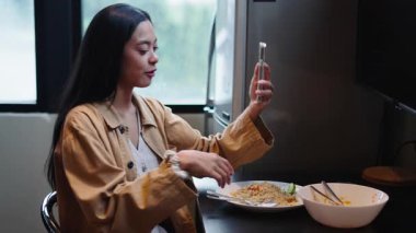 Mutfak masasında otururken aynı anda yemek yiyen mutlu genç bir kadın video araması yapıyor - 4K Yatay