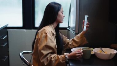 Genç Asyalı kadın sabah yemeğinin tadını çıkarırken videolu sohbet akıllı telefonlu masada oturuyor - 4K Yatay