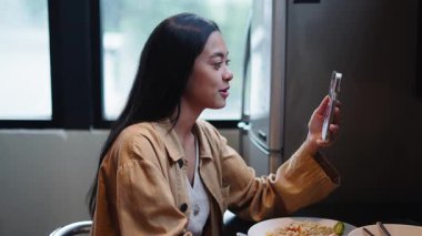 Genç bir kadın mutfakta akıllı telefon kullanarak öğle yemeğinden zevk alıyor - 4K Yatay