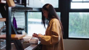 Pencere kenarında duran genç bir kadın ev yapımı yemekleri hazırlarken bir kasede malzemeleri karıştırıyor - 4K Yatay