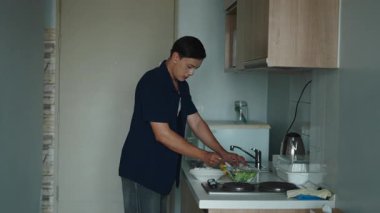 Genç yetişkin mutfak tezgahında dikilip dikkatlice taze sebzeleri kesiyor ve besleyici bir salata hazırlıyor - 4K Yatay