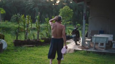 Güneydoğu Asyalı çiftçi elinde bir su bidonuyla evinin bahçesinde yürüyor - 4K Yatay