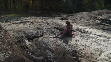 Dinlenen kadın yürüyüşçü kayalık arazide çağlayan şelalenin yanında oturuyor