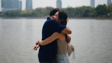 Sevgi dolu bir çift, deniz kenarında sıcak bir şekilde kucaklaşıyor, kentsel yeşil manzarada samimi bir bağ paylaşıyor.