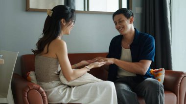 Asyalı çift rahatlıyor, el ele tutuşuyor, çağdaş oturma odasındaki kanepede otururken samimi bir şekilde sohbet ediyorlar.