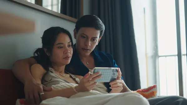 Genç çift yan yana dinleniyor, kanepede uzanırken akıllı telefon ekranlarını paylaşıyorlar.