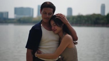 Romantik çift, arka planda bir şehir manzarası olan bir gölün kıyısında bir anı paylaşıyor.