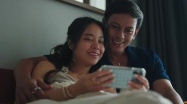 Güneydoğu Asyalı genç çift yatakta akıllı bir telefondan video izleyerek eğleniyor.