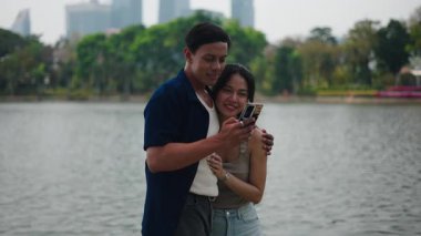 Romantik çift gölün kenarında eğleniyor, telefonlarıyla sosyal medyada geziniyorlar.