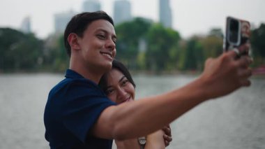 Gülümseyen milenyumlar, göl kenarında kentsel gökyüzü arkaplanı ile selfie anı yakalıyorlar.