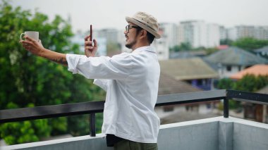 Kentin arka planında şehir balkonunda rahatlarken kahve fincanının fotoğrafını çeken yaratıcı serbest yazar