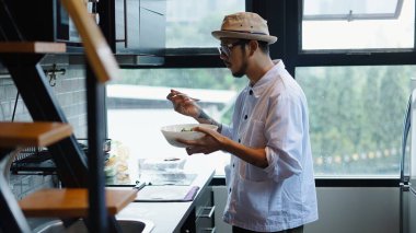 Aşçı şapkası ve gözlük takan profesyonel Asyalı şef büyük mutfak penceresinin yanında hazır mutfağı tadıyor.