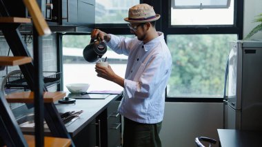 Şapkalı ve gözlüklü Asyalı bir adam modern bir mutfakta sıcak bir içecek hazırlıyor.