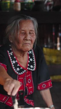 Geleneksel kıyafetler giymiş yaşlı bir kadın, FullHD dikey mutfağında, dumanı tüten bir tencerede yemek hazırlıyor.