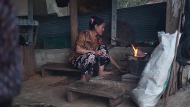 Yaşlı bir kadın ahşap bir platformda çıplak ayakla oturur ve kırsal bir mutfakta geleneksel bir soba ile ilgilenir.