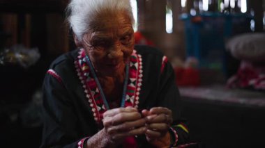 Yaşlı bir Asyalı kadın, geleneksel Tayland sahasında tekstil zanaatı üzerinde çalışmak için iğnenin ipliğine titizlikle iplik geçirir - 4K Yatay