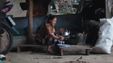 Yaşlı Taylandlı kadın, kırsal bir ortamda odun ateşinin yanındaki çaydanlığa çay yaprağı koyuyor. 4K Yatay.