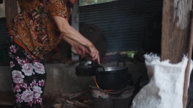 Asyalı bir kadın basit bir ortamda kırsal bir mutfakta geleneksel ev yapımı ahşap ateşiyle yemek hazırlıyor - 4K Yatay