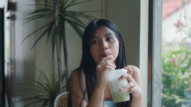 Bir kafede buzlu matcha latte yudumlayan genç bir kadın, pencereden dikkatlice dışarı bakıyor.