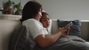 Asyalı anne ve kızı evdeki koltukta birlikte akıllı telefon kullanıyor - 4K Yatay
