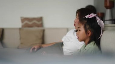 Asyalı anne ve kız oturma odasında televizyon izliyor ve ekranda bir şeyi işaret ediyor - 4K Yatay