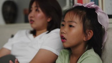 Asyalı anne-kız bir kanepede oturup televizyon seyrediyor ve farklı yüz ifadeleri sergiliyor - 4K Yatay