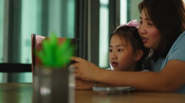 Bir anne, küçük kızına oturma odasındaki masada otururken tablet kullanmasında yardımcı olur - 4K Yatay