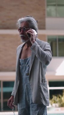 Uzun saçlı, güneş gözlüğü takan ve tulumlu bir Asyalı modern bir binanın havuzundan telefon görüşmesi yapıyor.