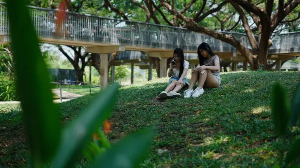 İki öğrenci parkta dinleniyor, akıllı telefonlarını yaya köprüsünün altında kullanıyorlar.