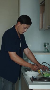 Genç yetişkin mutfak tezgahında dikiliyor taze sebzeleri dikkatlice kesiyor ve besleyici bir salata hazırlıyor - FullHD Dikey