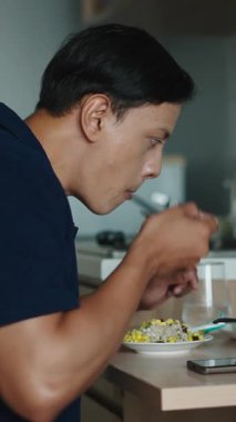 Genç bir adam modern mutfakta tek başına oturmuş su içerken taze salata yiyor akıllı telefonunun yanında.