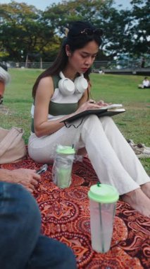 Şehir parkında güneşli bir günde çimlerin üzerinde oturan genç bir kadın manzara çiziyor.