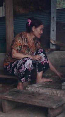 Yaşlı bir kadın ahşap bir platformda çıplak ayakla oturur kırsal bir mutfakta geleneksel bir soba üzerinde ateşe bakar.