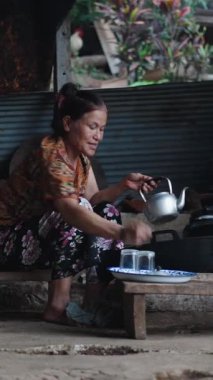 Yaşlı Taylandlı kadın, FullHD dikey bir kırsal alanda, odun ateşinin yanındaki çaydanlığın içine çay yaprakları koyuyor.