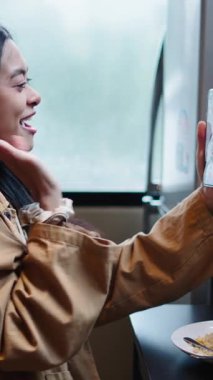 Mutlu genç Asyalı kadın yemek saatinde akıllı telefonuyla FullHD dikey mutfağında bir video görüşmesi yapıyor.