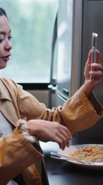 Mutfak masasında oturup yemek yerken aynı zamanda video görüşmesi yapan mutlu genç kadın FullHD Dikey