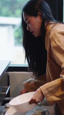 Uzun saçlı genç Asyalı kadın mutfak penceresinin yanında taze malzemeler pişirmeden önce mutfak gereçlerini hazırlıyor.