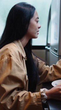 Genç Asyalı kadın sabah yemeğinin tadını çıkarırken videolu sohbet akıllı telefonlu masada dikey olarak oturuyor.