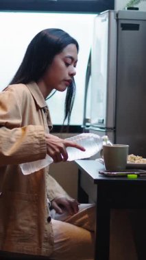 Genç Asyalı kadın masaya oturmadan önce şişeden büyük bardağa çok soğuk su dolduruyor.